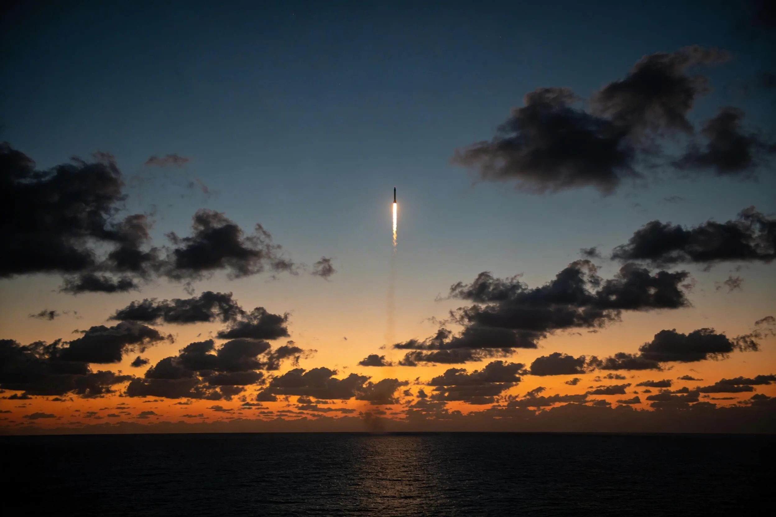 Raket stijgt bij zonsondergang op boven zee, tegen een oranje lucht met donkere wolken